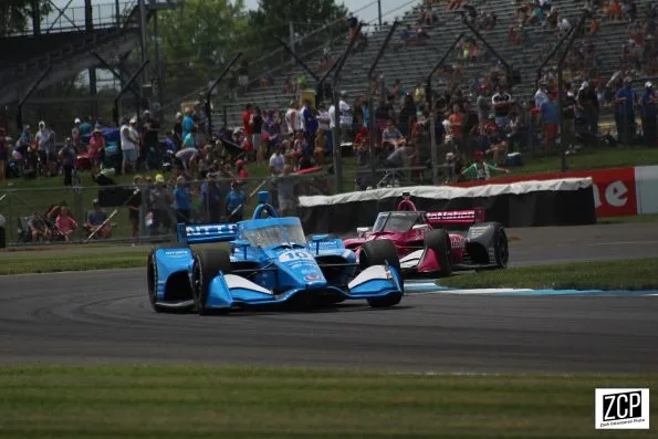 Álex Palou's blue NTT DATA Chip Ganassi Dallara-Honda, car 10, ahead of a pink AutoNation car on the Indianapolis Motor Speedway road course in 2021, grandstands behind.