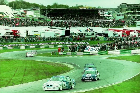A white and green Mobil Vauxhall Cavalier leading the two dark green Bailey ICS Ford Mondeos of Paul Radisich and Andy Rouse through McLaren corner at Brands Hatch in April 1994, with packed grandstands and transporters behind.