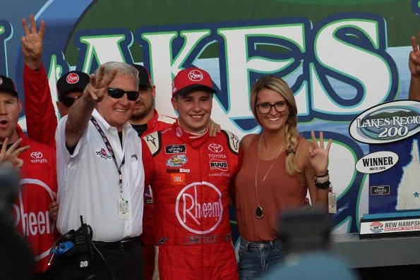 Christopher Bell in red Rheem overalls in victory lane at New Hampshire Motor Speedway in 2018, surrounded by team members, with a Lakes Region 200 winner sign behind him.