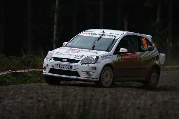 Elfyn Evans's white Ford Fiesta ST, car 118, on a wet forest gravel stage at Wales Rally GB in 2007, the family dealership web address gwyndafevansmotors.com printed across the bonnet.