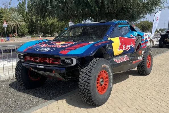 The blue and red Ford Raptor T1+ rally-raid car Mattias Ekström drives for M-Sport Ford, parked on paving with palm trees behind