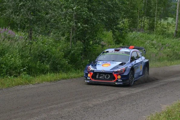 Thierry Neuville's blue Hyundai i20 Coupe WRC on the gravel of the Saalahti stage at Rally Finland in 2017, kicking up dust with fireweed on the bank behind.